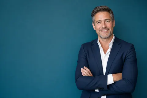 A real estate broker in a navy blazer with arms folded against a teal wall