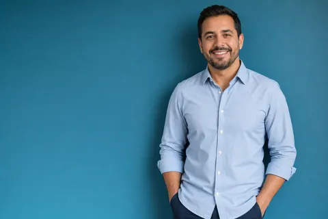 A real estate agent in a light blue shirt with hands in pockets against a teal wall