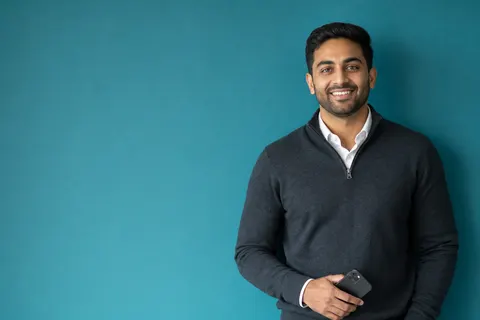 A newly licensed real estate agent in a charcoal sweater holding a phone against a teal wall