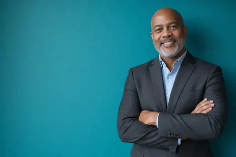A real estate broker in a charcoal suit jacket with arms folded against a teal wall